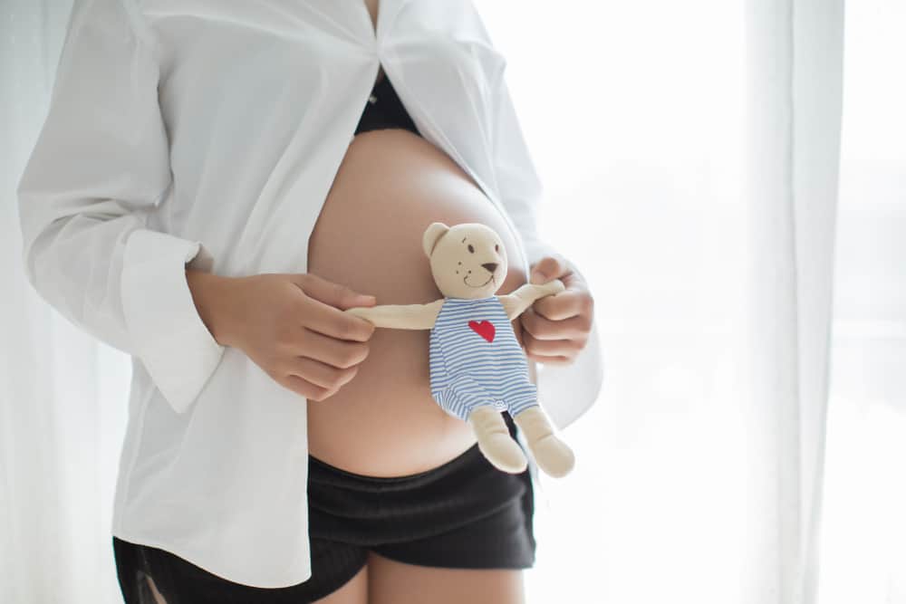 Pregnant woman smiling and touching her belly while standing near a window with morning light